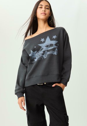 Felpa grigia, scollata sulle spalle, con una stampa grafica di un'auto e stelle. Presenta maniche lunghe e orlo a coste, abbinata a pantaloni cargo neri.