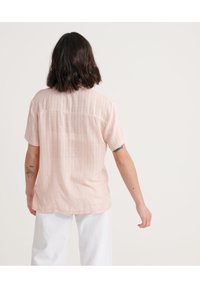 Camicia rosa chiaro a maniche corte con cucitura posteriore e sottili righe verticali, realizzata in tessuto morbido, abbinata a pantaloni bianchi.