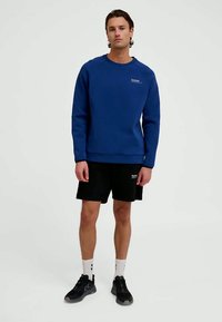 Zils džemperis ar ribotām aprocēm un apakšmalu, apaļa kakla izgriezums, valkāts virs melnajām īsām biksēm. Redzami melni sporta apavi un baltas zeķes.