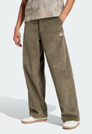 Osoba nosząca luźne oliwkowozielone spodnie sztruksowe z logo Adidas na kieszeni, w połączeniu z białymi trampkami i casualową koszulą.