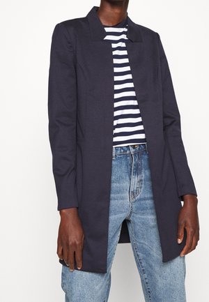 Person iført en mørk marineblå lang blazer over en sort-hvid stribet skjorte og lyseblå højtaljede jeans, stående mod en hvid baggrund.