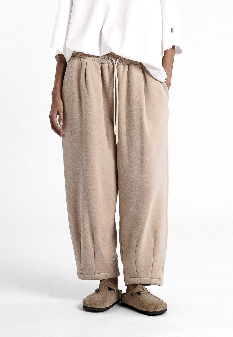 Pantalons de survêtement beiges, ample, avec une taille élastique et un cordon de serrage. Tissu doux et poignets resserrés. Associés à des chaussons beiges.