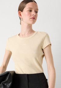 Lys beige ribbet t-skjorte med korte ermer og rund hals; med en subtil brodert detalj foran.