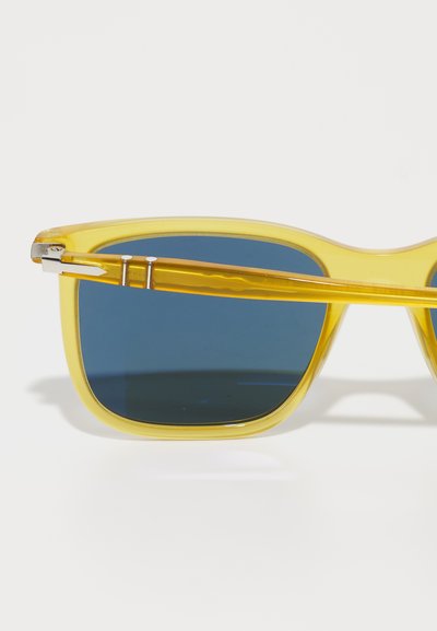 Óculos de sol de plástico amarelo com lentes quadradas, apresentando lentes com tonalidade azul, metal nas dobradiças e um design elegante nas hastes.