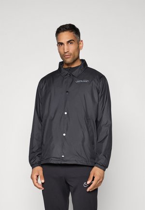 Black nylon jacket with snap buttons and a collar, featuring a logo on the chest and elastic cuffs. Worn over black pants.
