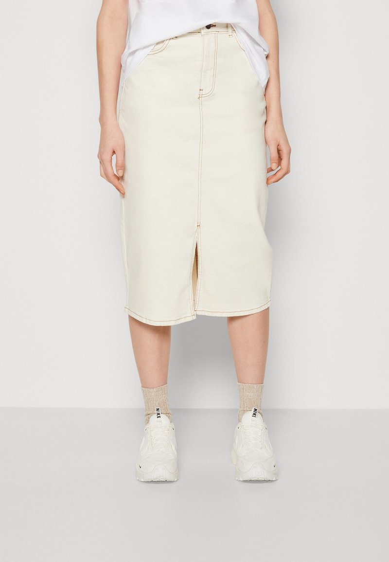 Jupe midi en denim beige avec une fente avant, des détails de coutures visibles et un ourlet légèrement arrondi. Associée à des baskets blanches et des chaussettes beiges.