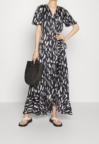 Robe portefeuille noire et blanche à motifs, avec des manches courtes bouffantes et un ourlet volanté, assortie à un sac tressé noir et des sandales blanches.