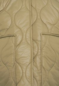 Μπεζ καπιτονέ μπουφάν με κυματιστό σχέδιο, δύο μπροστινές τσέπες και λεία, γυαλιστερή υφή. Ραφές κατά μήκος των άκρων.