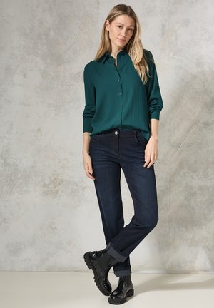 Chemise bleue canard à manches longues avec des plis verticaux, assortie à un pantalon en denim sombre et à des bottes noires à semelles épaisses. Tissu lisse et détails de manches retroussées.