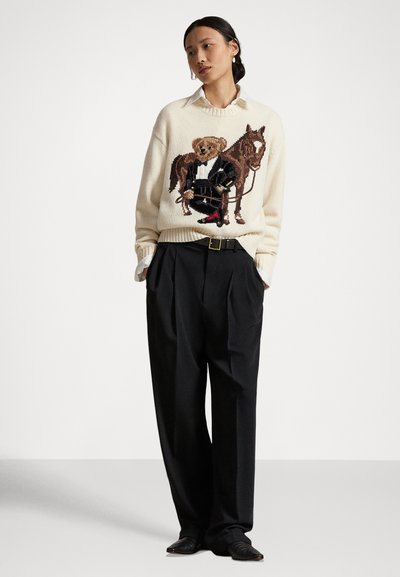 Kobieta w kremowym swetrze z grafiką misia w smokingu i konia, zestawionym z czarnymi plisowanymi spodniami i czarnym paskiem.