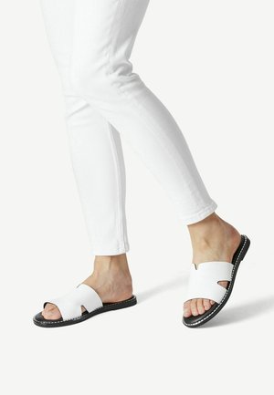 Sandales blanches avec un design découpé, semelle noire et couture blanche. Portées avec un pantalon blanc ajusté, montrant un détail sur les orteils et le pied.