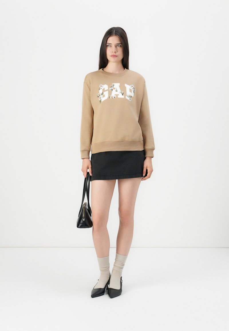 Joven de pie, con sudadera floral color crema de GAP, falda mini negra, calcetines beige con tacones negros, sosteniendo un bolso negro frente a un fondo blanco.
