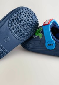 Des chaussures en caoutchouc bleues avec une semelle texturée, une sangle réglable et un détail de personnage de dessin animé vert sur le côté.