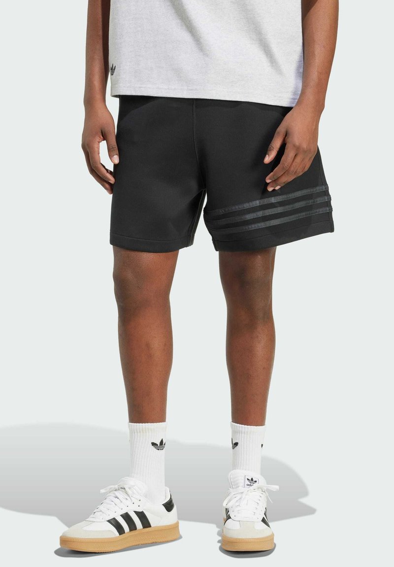 Černé sportovní šortky se třemi vodorovnými tmavými pruhy po straně. V kombinaci s bílými teniskami s černými akcenty a bílými ponožkami.