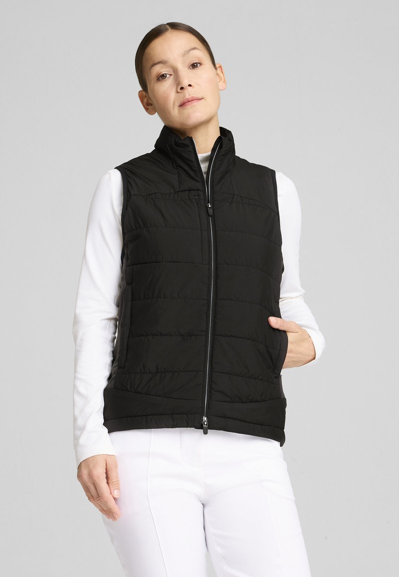 Chaleco negro tipo acolchado con cuello alto y cierre de cremallera, con un diseño acolchado y sin mangas, usado sobre una camiseta de manga larga blanca.