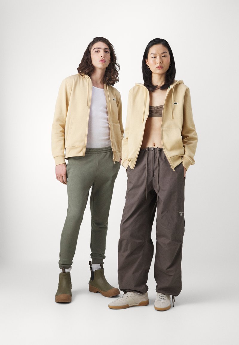 Twee personen staan samen. De één draagt een lichtgele hoodie met rits over een witte tanktop, olijfgroene broek en groene laarzen. De ander draagt een beige hoodie over een bruine bralette, losse grijze cargobroek met trekkoorden en witte schoenen.