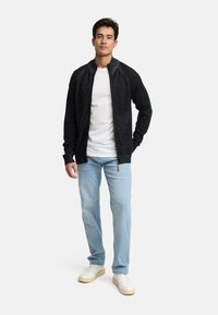Sort strikjakke med lynlås, struktureret stof, tætsiddende ærmer og en opretstående krave. Kombineret med lysblå jeans og hvide sneakers.