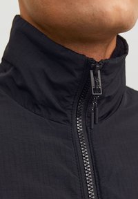 Svart zip-up jakke med stående krage, med teksturert overflate og kontrasterende glidelåsdetaljer. Stoffet ser lett og slitesterkt ut.