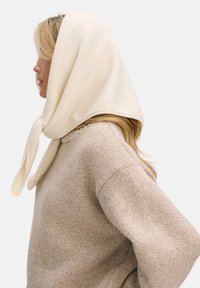 Jasnobeżowy dzianinowy sweter o szerokich rękawach, zestawiony z miękkim, kremowym szalikiem zawiązanym pod brodą, prezentujący fakturowane wykończenie.