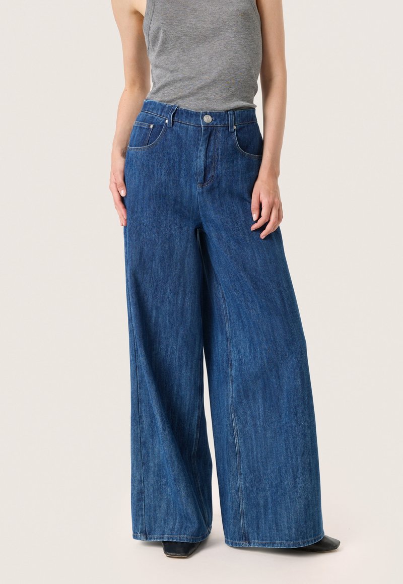 Modré džíny s širokými nohavicemi a vysokým pasem, s knoflíkovým zapínáním, pěti kapsami a hladkým povrchem. V kombinaci se šedým tílkem.