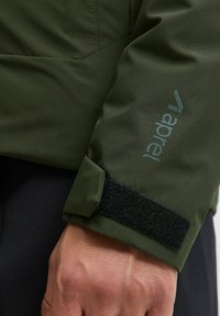 Tessuto della giacca verde oliva con una texture liscia; presenta una regolazione del polsino in Velcro e un sottile logo azzurro vicino al polso.