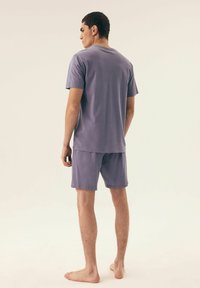 Lila Baumwoll-T-Shirt und passende Shorts. Das T-Shirt hat einen Rundhalsausschnitt, und beide Teile haben eine glatte, weiche Textur. Das Model steht barfuß und ist seitlich ausgerichtet.