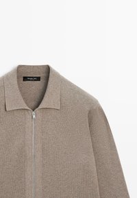 Beige gestrickte Jacke mit Reißverschluss vorne und kurzem Kragen, mit glatter Textur und geradem Schnitt. Markenetikett ist innen am Kragen sichtbar.