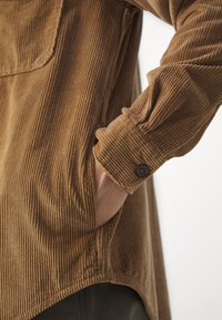 Brun fløjlsbluse med lodrette riller, der har en lomme, manchetknapdetalje og afslappet pasform ved kanten.