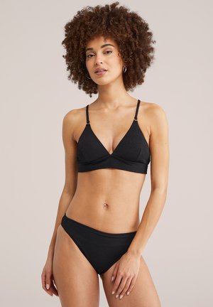 Vrouw met krullend haar die een zwart textuurbikini draagt met verstelbare bandjes, tegen een effen beige achtergrond.
