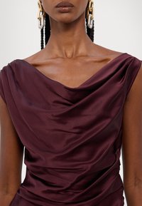 Robe en satin bordeaux avec un décolleté drapé et des détails aux épaules. Le tissu a une texture lisse avec un éclat subtil. Boucles d'oreilles en chaîne dorée et argentée.