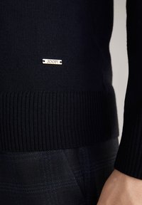 Marineblauer Strickpullover mit geripptem Saum, verziert mit einem kleinen silbernen Etikett mit der Aufschrift "JOOP!" an der unteren Seite, kombiniert mit dunkelkarierter Hose.