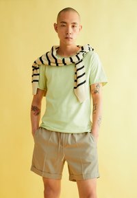 Hellgrünes T-Shirt mit kurzen Ärmeln, kombiniert mit beigen Shorts. Ein gestreifter Pullover liegt über den Schultern. Dazu sichtbare Tattoos.