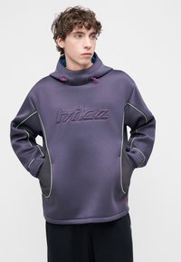 Lilla pusa kõrge kraega, millel on rohelised aktsendid, tikitud logo ja võrkkangast külgpaneelid. Kapuutsil on roosad nöörid.