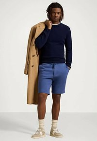 Man som bär en marinblå tröja, blå dragkedjshorts, beige sneakers och krämfärgade strumpor, hållande en kamelrock över ena axeln.