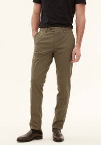 Chinos in cotone verde oliva con vestibilità dritta, dotati di chiusura a bottone, tasche laterali e una texture morbida. Abbinati a scarpe slip-on marroni.