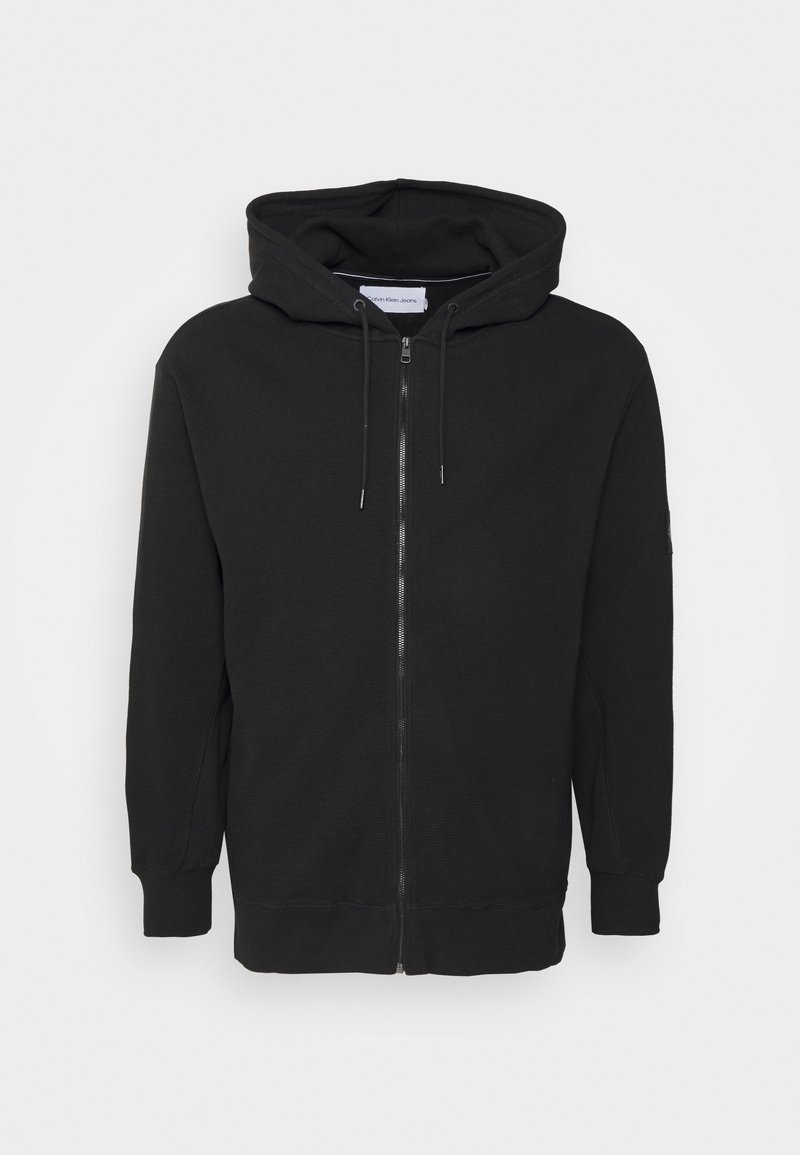 Sweat à capuche zippé noir avec cordons, poignets et bas côtelés, présenté sur un fond blanc uni.