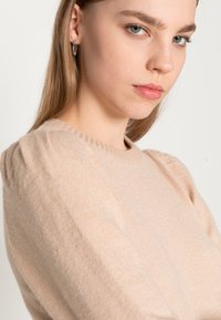 Svijetlosmeđa pulover s okruglim izrezom, s napuhanim rukavima i mekom teksturom. Model ima dugu kosu i srebrne kružne naušnice.