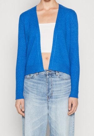 Frau trägt eine leuchtend blaue, offene Strickjacke, ein weißes, bauchfreies Tanktop und hoch taillierte, hellblaue Jeans.