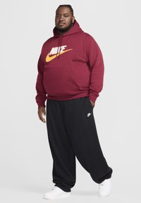 Sudadera Nike color marrón con logotipo blanco y amarillo, combinada con pantalones negros de corte holgado y zapatillas blancas. Ropa deportiva casual.