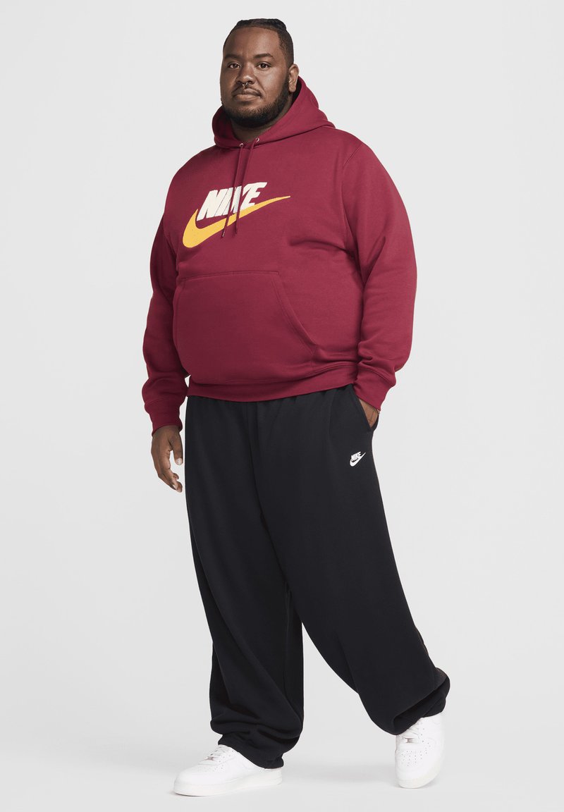 Sudadera Nike color marrón con logotipo blanco y amarillo, combinada con pantalones negros de corte holgado y zapatillas blancas. Ropa deportiva casual.