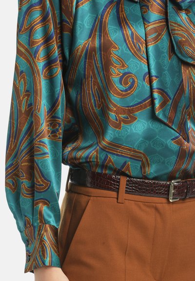 Satin blouse in turquoise met ingewikkelde bruine en blauwe patronen. Gecombineerd met bruine broeken en een donkere riem met krokodillenstructuur.