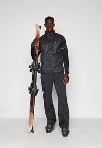 Muškarac u crnoj skijaškoj opremi drži jedan par skija uspravno, stojeći na sivoj podlozi nasuprot bijeloj pozadini.