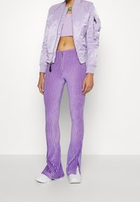 Světle fialová bomber bunda s lesklým povrchem, spárovaná s vertikálně pruhovanými fialovými rozšířenými kalhotami a bílými teniskami.