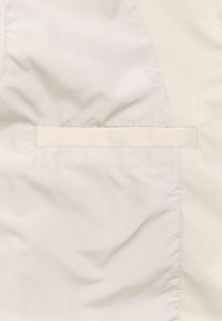 Lichtbeige pufferjack met een gladde textuur. Kenmerkt zich door een rechthoekige zakdetail langs de rand en een over het geheel zachte, gewatteerde uitstraling.