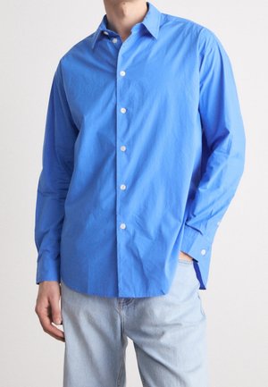 Mann trägt ein langärmeliges, hellblaues Hemd mit Knöpfen und helle blaue Jeans, steht mit einer Hand in der Tasche vor einfarbigem Hintergrund.