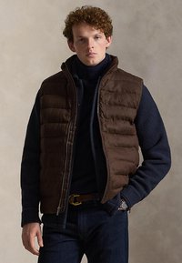 Młody mężczyzna z kręconymi włosami, ubrany w ciemnobrązową pikowaną kamizelkę na granatowym polarze, niebieskich dżinsach i zegarku na nadgarstku, stojący na jednolitym tle.