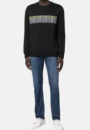 Felpa nera con colletto a girocollo, che presenta la scritta in contrasto "BOSS" in giallo e bianco sul petto. Indossata con jeans blu e sneaker bianche.