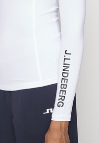 White long-sleeve shirt with a black "J.LINDEBERG" print on the sleeve, paired with navy ribbed pants featuring a white logo accent.