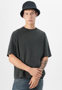 Camiseta gris de gran tamaño con escote redondo, mangas cortas y costuras visibles, combinada con un sombrero tipo cubo negro.
