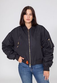 Czarna kurtka bomberka wykonana z gładkiego, lekkiego materiału; posiada złote zamki błyskawiczne, ściągacze przy mankietach oraz puszyste rękawy z metką na dolnym wykończeniu.